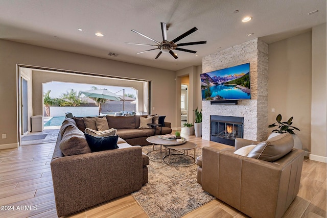 living room with a stone fireplace, a textured ceiling, ceiling fan, and light hardwood / wood-style flooring