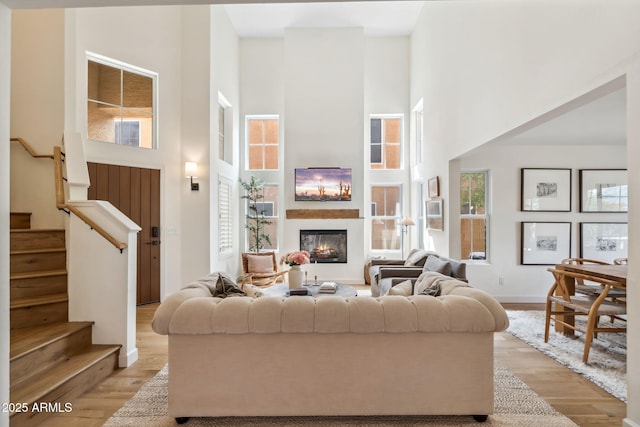 living room with a high ceiling, a warm lit fireplace, light wood-type flooring, and stairs