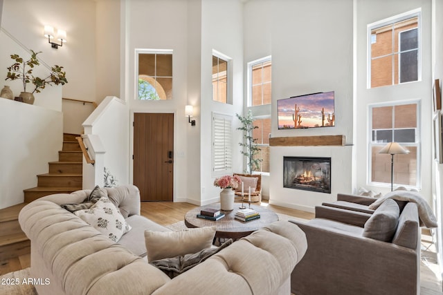 living area featuring a towering ceiling, light wood finished floors, a glass covered fireplace, stairway, and baseboards