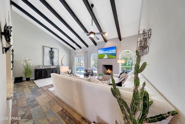 living area featuring beamed ceiling, a warm lit fireplace, a ceiling fan, and high vaulted ceiling