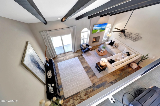 living area featuring ceiling fan, beamed ceiling, a warm lit fireplace, and wood finished floors