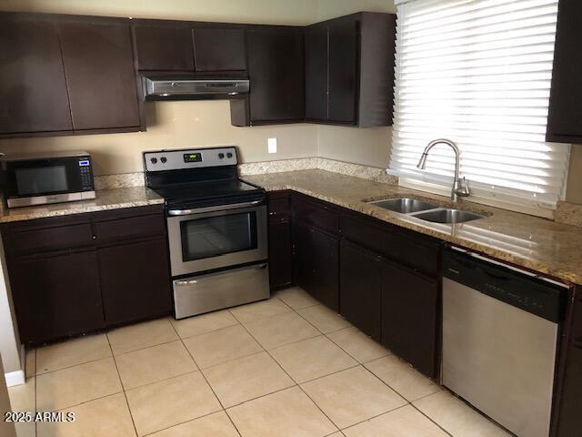 kitchen with under cabinet range hood, light tile patterned floors, appliances with stainless steel finishes, stone countertops, and a sink