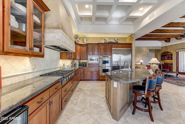 kitchen featuring built in appliances, a sink, beam ceiling, dark stone countertops, and a kitchen bar