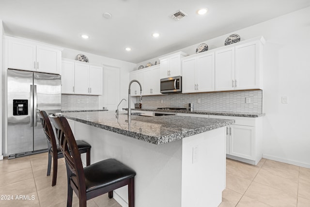 kitchen featuring white cabinetry, stainless steel appliances, a center island with sink, and dark stone counters