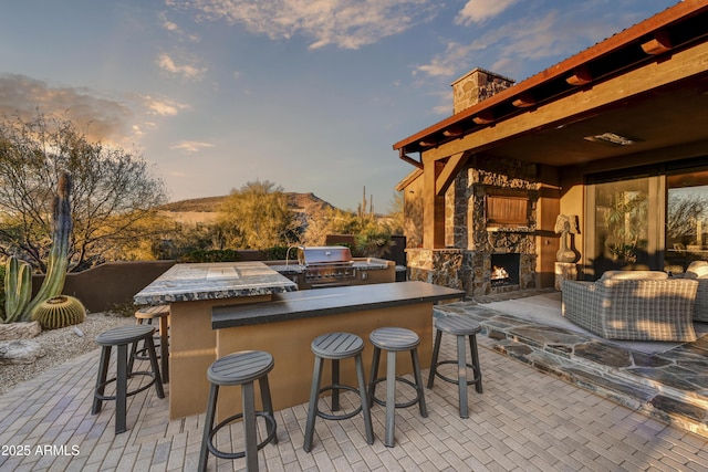 patio terrace at dusk with exterior bar, an outdoor stone fireplace, exterior kitchen, a mountain view, and a grill