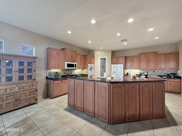 kitchen featuring backsplash, dark stone counters, white appliances, a center island with sink, and light tile patterned floors