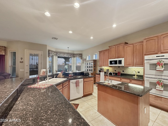 kitchen featuring white appliances, a kitchen island with sink, sink, hanging light fixtures, and dark stone countertops