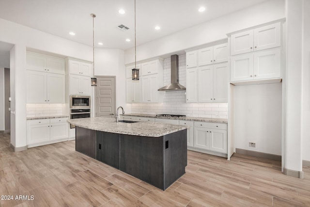 kitchen with appliances with stainless steel finishes, wall chimney exhaust hood, a center island with sink, white cabinets, and hanging light fixtures