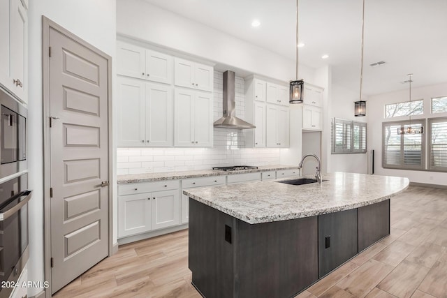 kitchen featuring wall chimney range hood, sink, an island with sink, decorative light fixtures, and white cabinetry