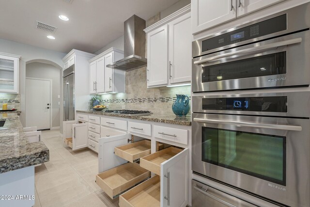 kitchen with wall chimney range hood, stainless steel appliances, light stone countertops, decorative backsplash, and white cabinets