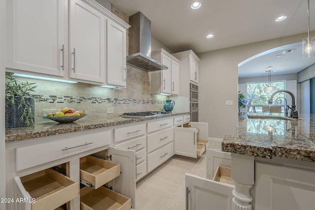 kitchen featuring wall chimney range hood, white cabinetry, stainless steel appliances, light stone countertops, and decorative light fixtures