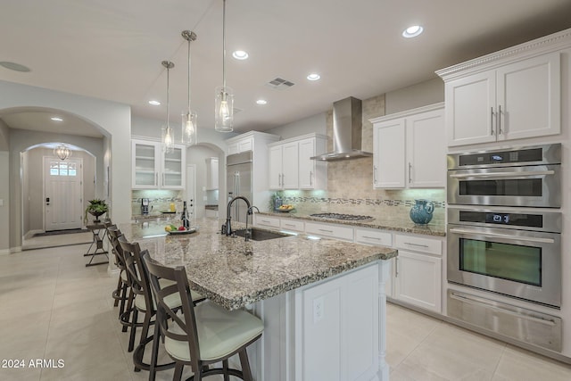 kitchen featuring wall chimney range hood, white cabinetry, stainless steel appliances, decorative light fixtures, and a large island with sink