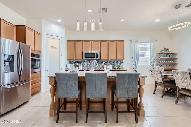 kitchen featuring a kitchen island with sink, backsplash, stainless steel appliances, light stone countertops, and decorative light fixtures