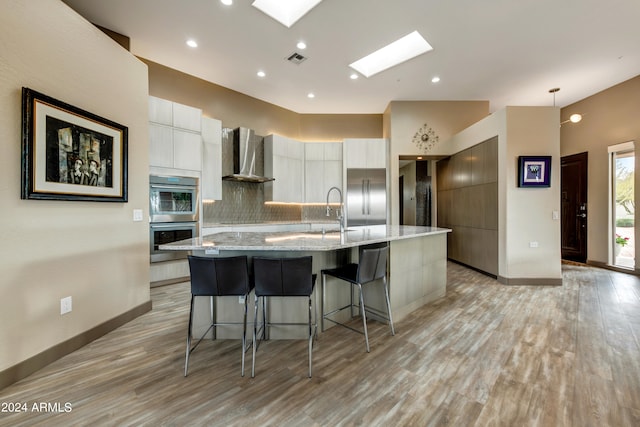 kitchen with appliances with stainless steel finishes, light hardwood / wood-style floors, wall chimney range hood, an island with sink, and a skylight