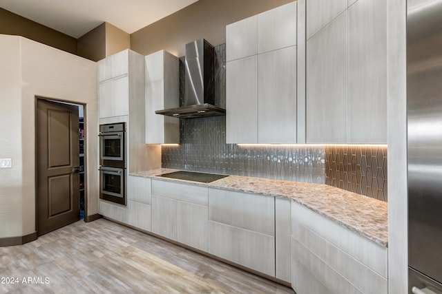 kitchen featuring light stone counters, double oven, wall chimney range hood, backsplash, and light hardwood / wood-style flooring