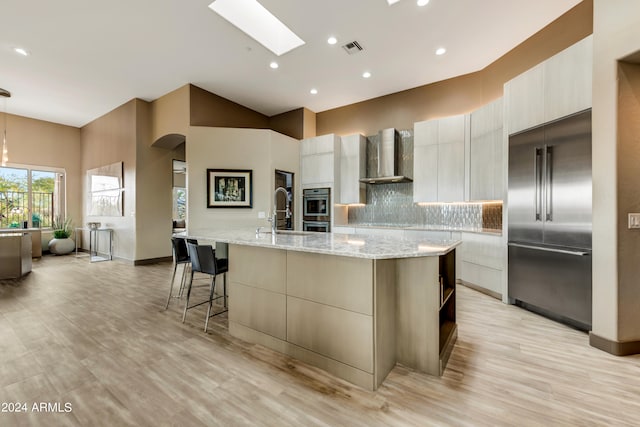 kitchen with a skylight, an island with sink, appliances with stainless steel finishes, and wall chimney range hood