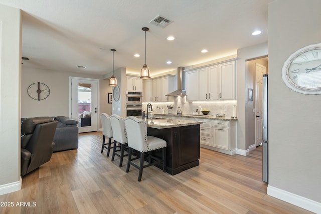 kitchen with a breakfast bar, white cabinets, light stone countertops, a center island with sink, and wall chimney exhaust hood