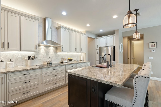 kitchen with hanging light fixtures, stainless steel refrigerator with ice dispenser, black electric stovetop, an island with sink, and wall chimney exhaust hood