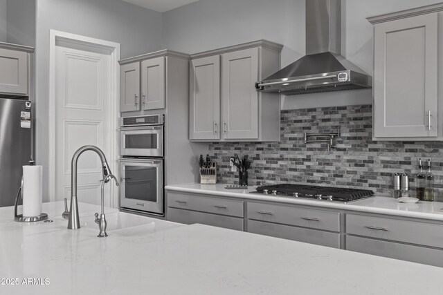 kitchen featuring a sink, gray cabinetry, appliances with stainless steel finishes, wall chimney range hood, and tasteful backsplash