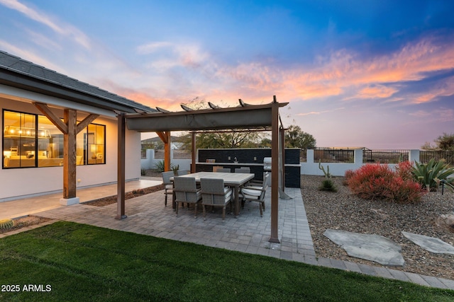 patio terrace at dusk featuring a lawn, fence, and exterior kitchen