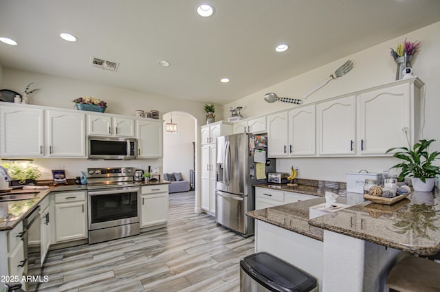 kitchen with kitchen peninsula, dark stone countertops, appliances with stainless steel finishes, light hardwood / wood-style floors, and white cabinetry