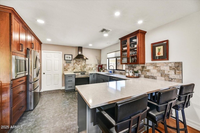 kitchen with backsplash, appliances with stainless steel finishes, a peninsula, glass insert cabinets, and wall chimney exhaust hood