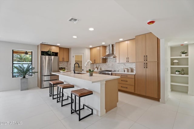 kitchen with sink, wall chimney exhaust hood, stove, stainless steel fridge, and a center island with sink