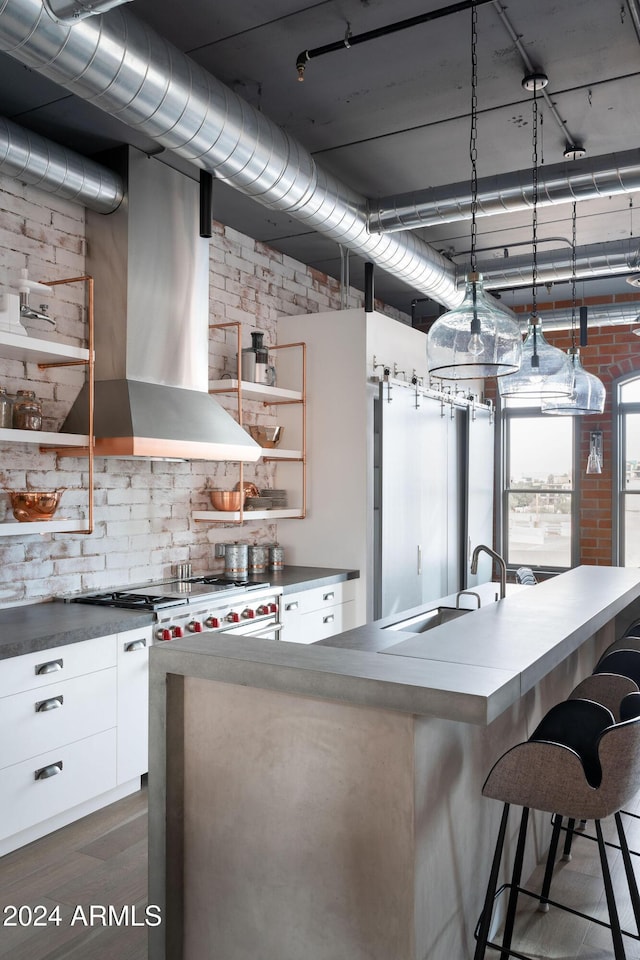 kitchen featuring wall chimney exhaust hood, brick wall, sink, white cabinets, and hanging light fixtures