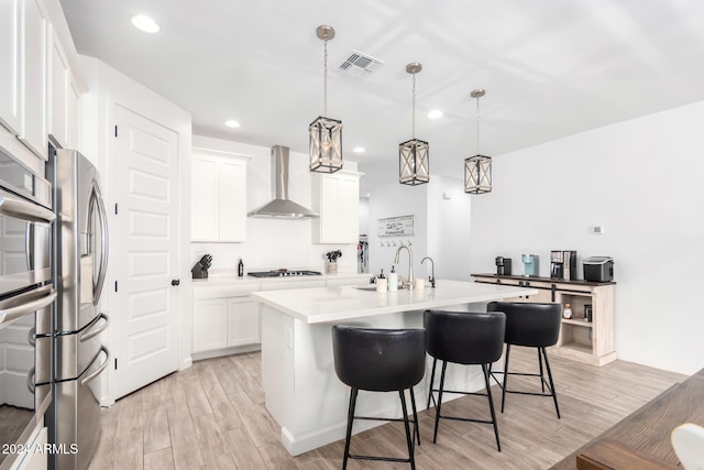 kitchen with light hardwood / wood-style floors, white cabinetry, wall chimney exhaust hood, an island with sink, and pendant lighting