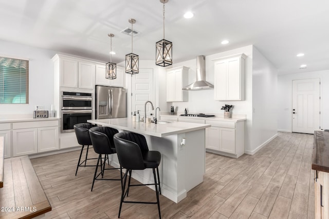 kitchen with a kitchen island with sink, wall chimney range hood, appliances with stainless steel finishes, hanging light fixtures, and light hardwood / wood-style flooring