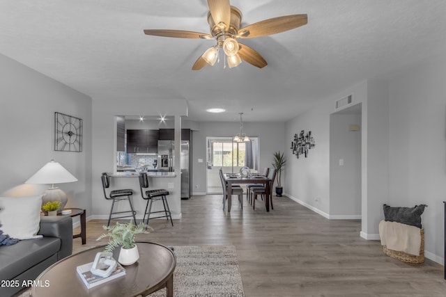 living room with visible vents, a textured ceiling, wood finished floors, baseboards, and ceiling fan with notable chandelier