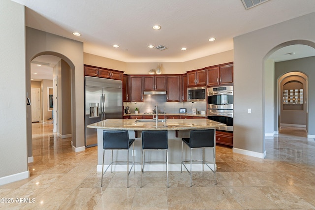 kitchen with built in appliances, a breakfast bar area, light stone countertops, and an island with sink