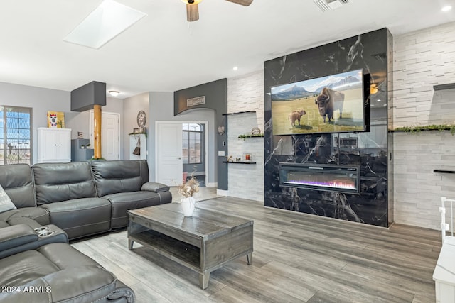 living room with light hardwood / wood-style flooring, ceiling fan, a skylight, and a stone fireplace