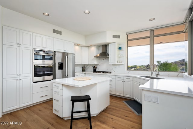 kitchen with wall chimney range hood, sink, white cabinetry, stainless steel appliances, and a center island