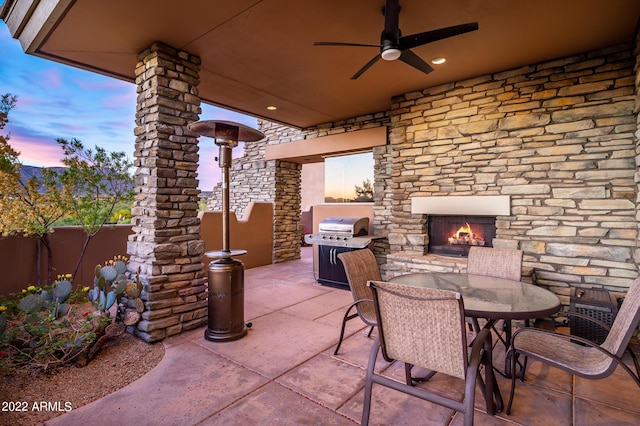 patio terrace at dusk featuring ceiling fan, area for grilling, and an outdoor stone fireplace