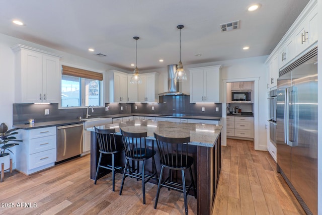 kitchen with pendant lighting, built in appliances, white cabinets, a kitchen island, and wall chimney exhaust hood