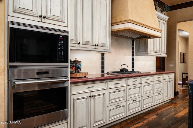 kitchen featuring appliances with stainless steel finishes, custom range hood, dark countertops, dark wood-style flooring, and tasteful backsplash