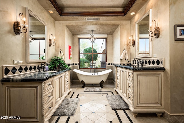 full bathroom with inlaid floor details, ornamental molding, a freestanding tub, two vanities, and a chandelier