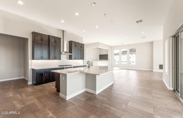 kitchen with visible vents, light countertops, wall chimney range hood, tasteful backsplash, and an island with sink