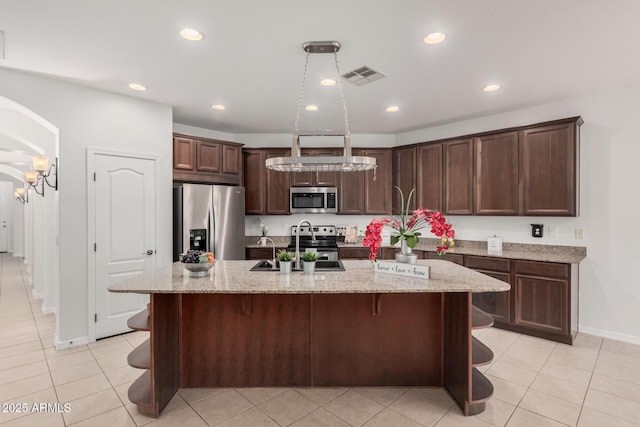 kitchen with arched walkways, dark brown cabinetry, stainless steel appliances, visible vents, and an island with sink