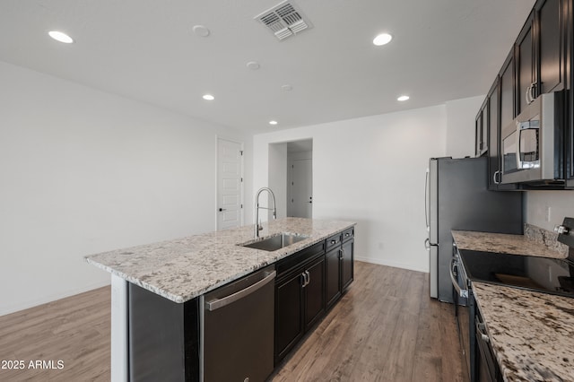 kitchen with an island with sink, sink, light stone counters, stainless steel appliances, and light wood-type flooring