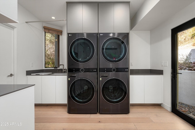 clothes washing area featuring stacked washer / drying machine, sink, light hardwood / wood-style floors, and cabinets