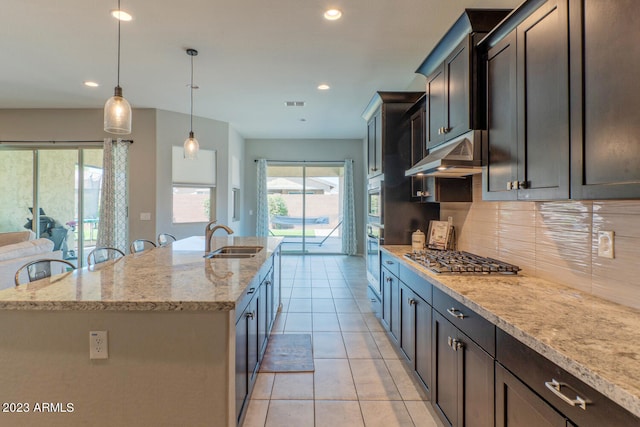 kitchen with sink, stainless steel appliances, light stone counters, hanging light fixtures, and a center island with sink