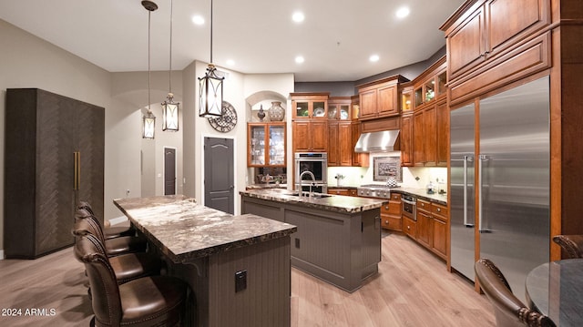 kitchen featuring dark stone counters, stainless steel appliances, a kitchen island with sink, exhaust hood, and hanging light fixtures