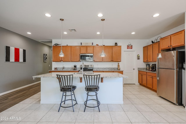 kitchen with stainless steel appliances, light stone counters, hanging light fixtures, and a kitchen island with sink