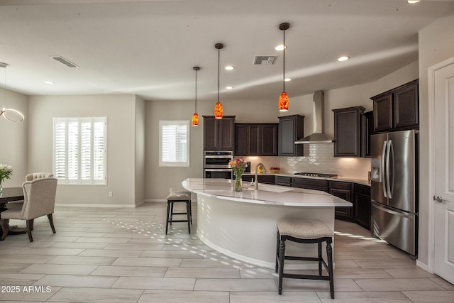 kitchen with wall chimney exhaust hood, a breakfast bar area, decorative light fixtures, stainless steel appliances, and a kitchen island with sink