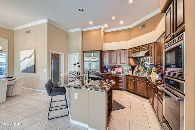 kitchen with dark stone countertops, a breakfast bar area, a kitchen island with sink, built in appliances, and light tile patterned floors