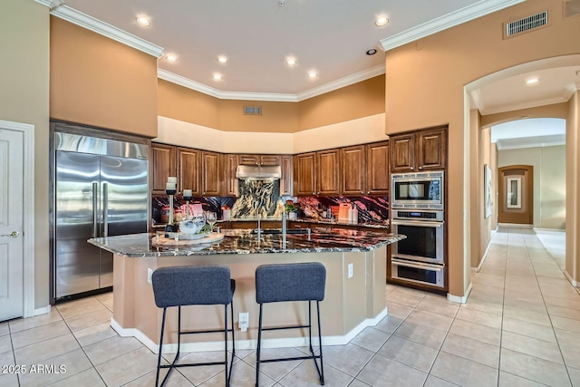 kitchen with light tile patterned floors, built in appliances, a kitchen island with sink, and dark stone countertops