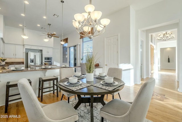 dining space with visible vents, light wood-style flooring, a high ceiling, and ceiling fan with notable chandelier