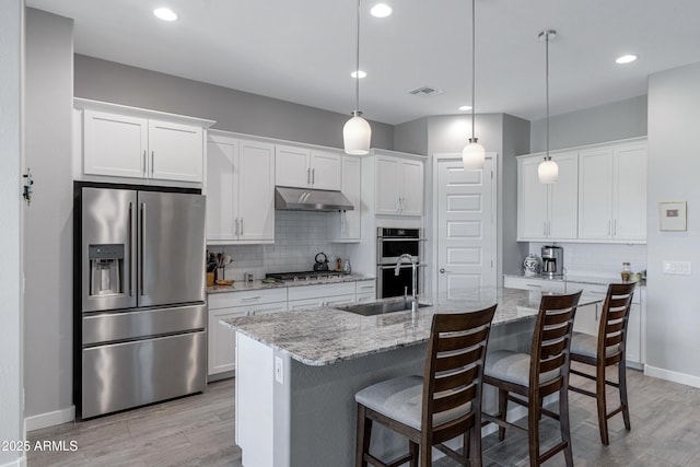 kitchen with light wood finished floors, under cabinet range hood, appliances with stainless steel finishes, white cabinets, and a sink